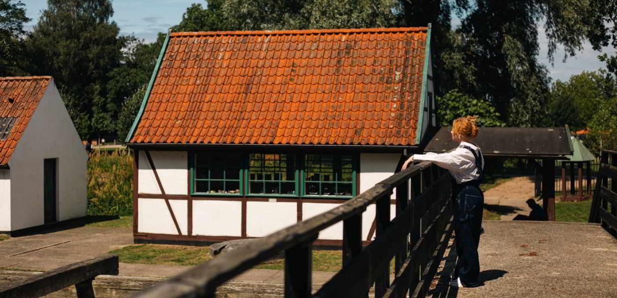 En ung kvinde står på en bro og kigger på et lille bindingsværkshus på Krudtværksmuseet